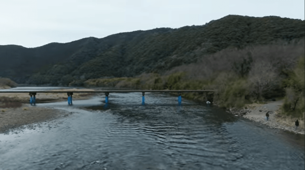 Bridge spans a calm river with blue supports, set in a forested valley with hills in the background; a few people stand on the right bank.