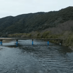 Bridge spans a calm river with blue supports, set in a forested valley with hills in the background; a few people stand on the right bank.