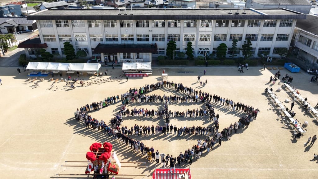 Aerial view of a schoolyard with students standing in a large peace-sign formation, tents and a stage visible nearby.