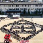 Aerial view of a schoolyard with students standing in a large peace-sign formation, tents and a stage visible nearby.