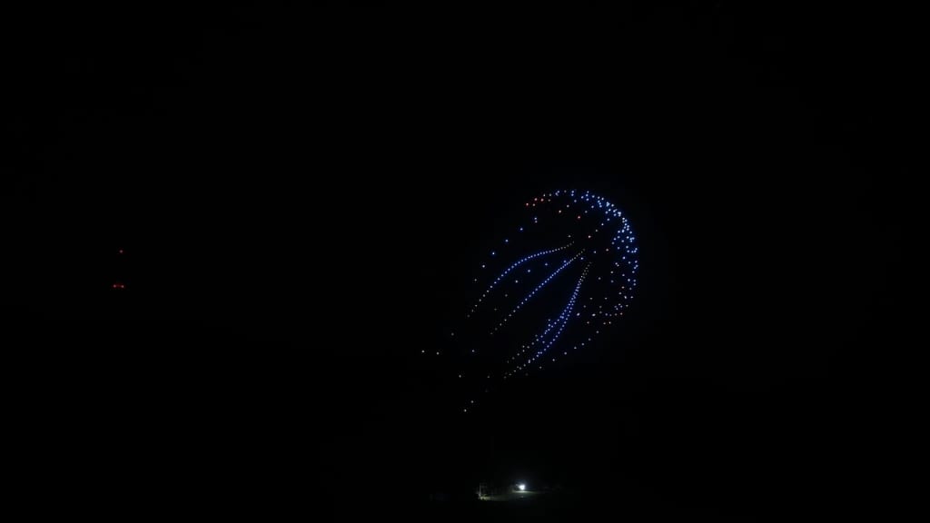 Blue fireworks form a spiral of dotted trails in the dark sky, with a small lit area on the ground below.