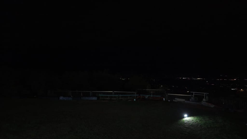 Night scene of a grassy area at night with a single bright ground light illuminating a patch of grass and a railing along the edge, distant city lights on the horizon.