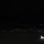 Night scene of a grassy area at night with a single bright ground light illuminating a patch of grass and a railing along the edge, distant city lights on the horizon.