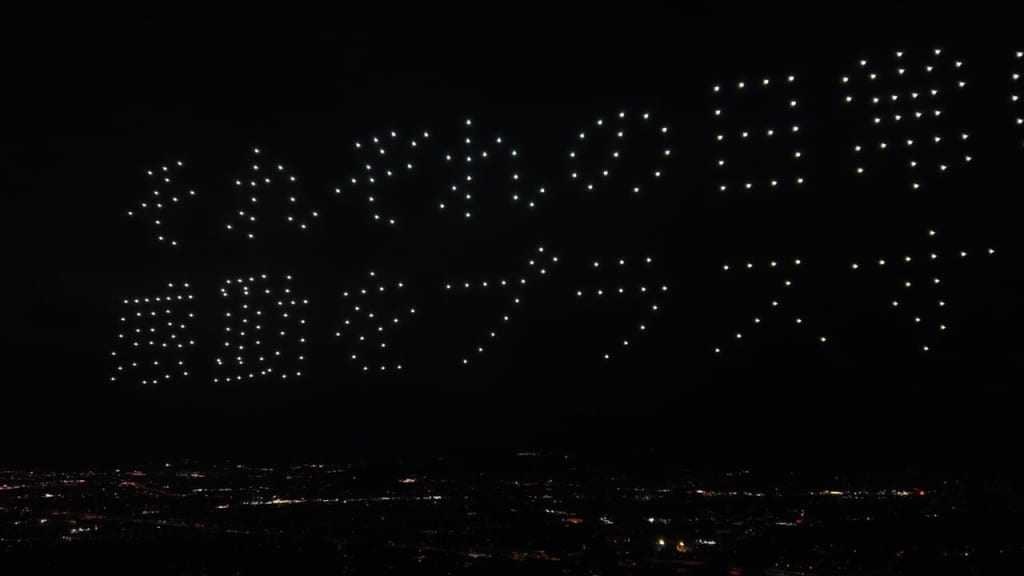 Aerial drone light display forms a message in the dark night sky over a cityscape with scattered streetlights below.