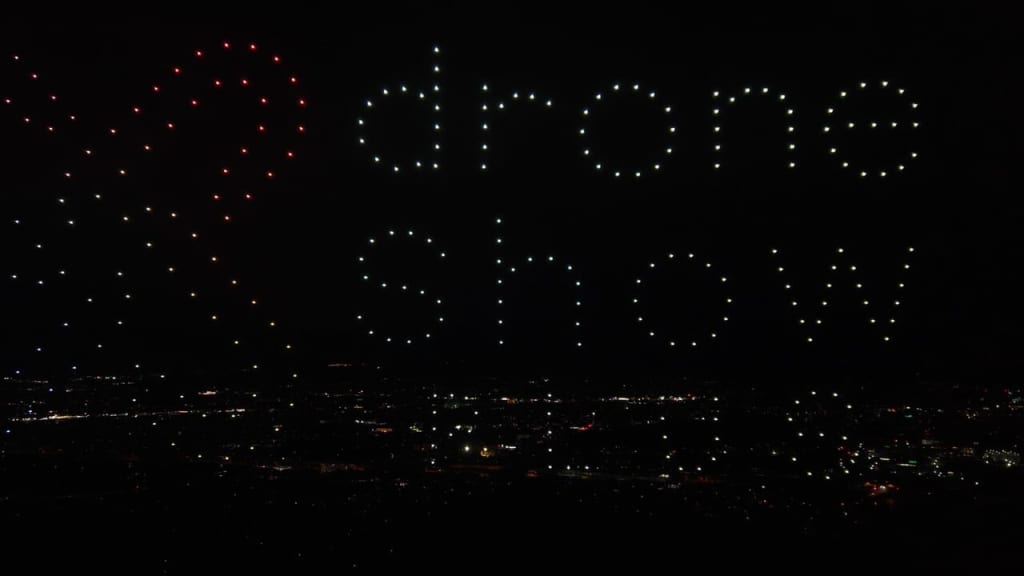 Drone light show over a city at night, forming a red heart on the left and white letters in the sky to the right.