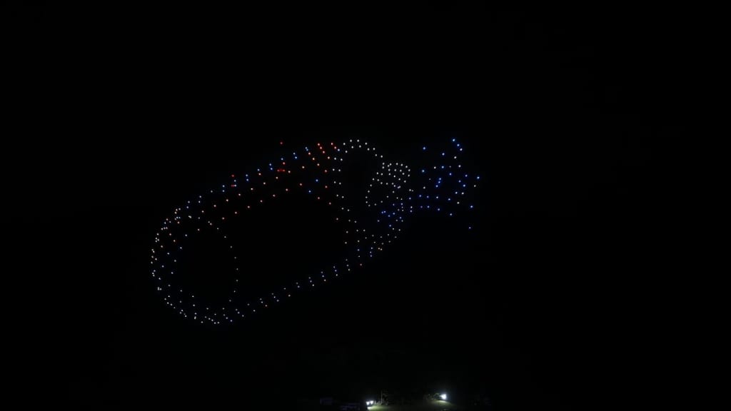 Dark sky with a large drone-light display forming an oval loop of red, white, and blue lights; a blue tail of lights trails to the right above a small lit area on the ground.