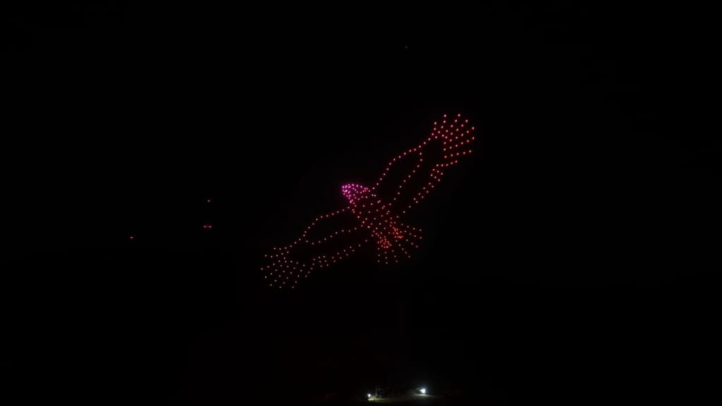 Red LED drone light formations arranged in a double-wing shape glowing in the night sky above a dark ground.