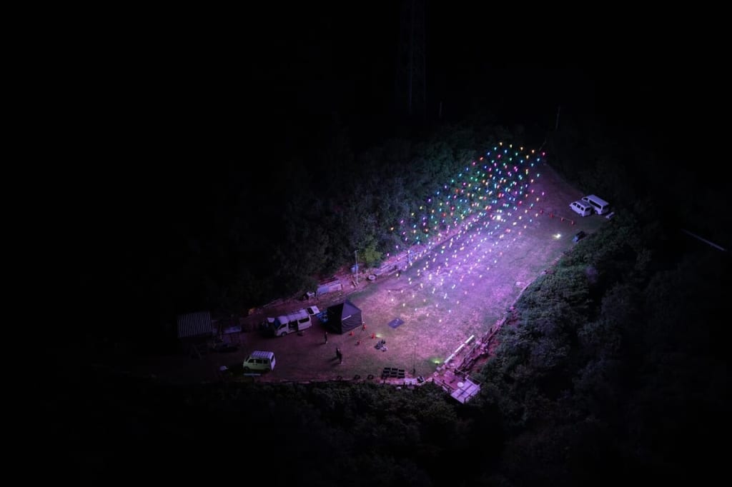 Nighttime campsite with a diagonal fan of colorful string lights over a grassy area, vehicles and tents nearby.