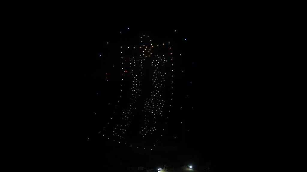Night sky illuminated by a large dotted-firework pattern with small colorful sparks around; ground lights faintly visible below.