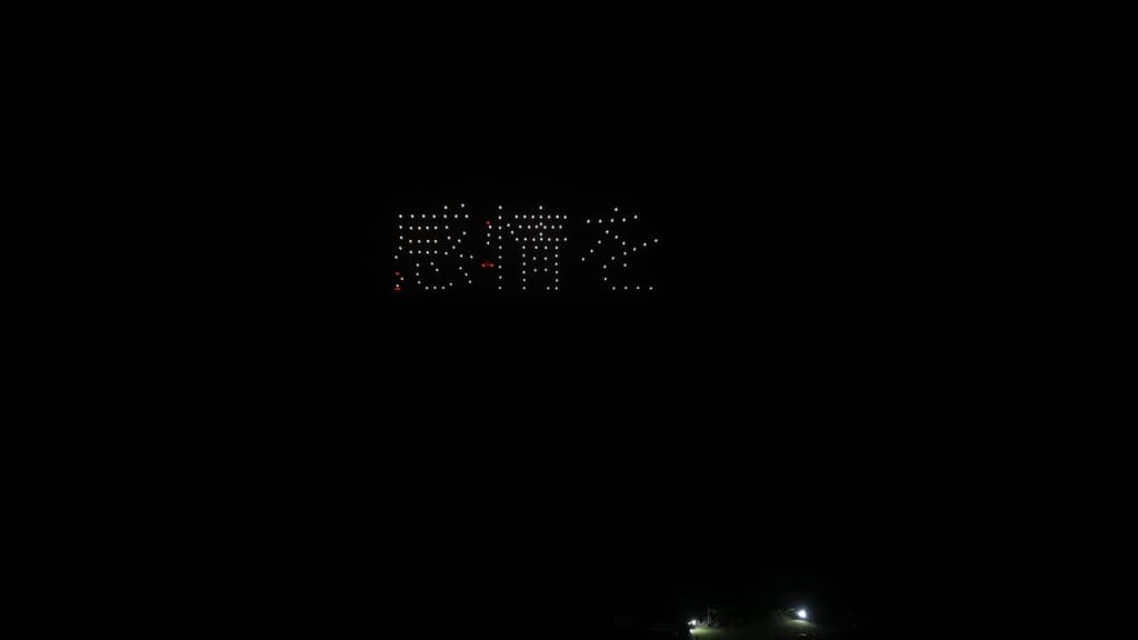 Distant LED sign in a dark night, formed by small white lights with a few red accents, set high on a hillside. A pair of bright ground lights is visible below.