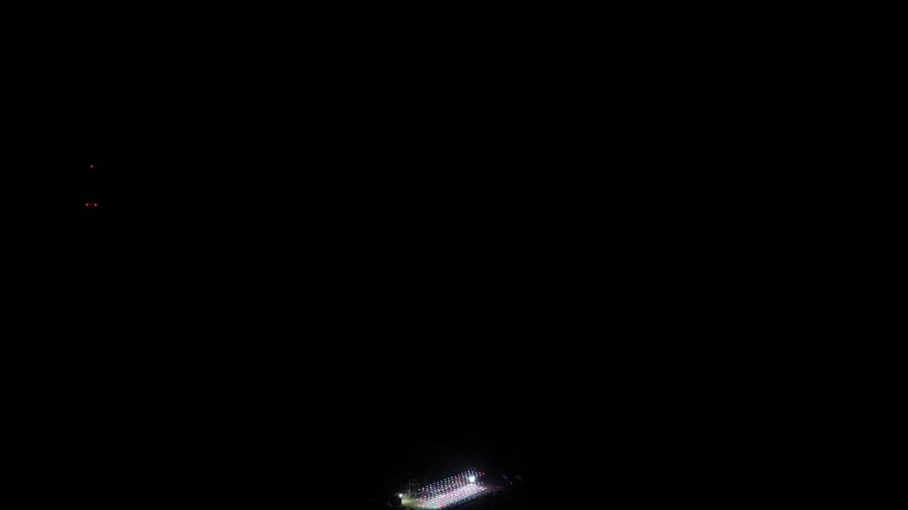 Night scene with a small, brightly lit stadium or building near the bottom center, surrounded by darkness.